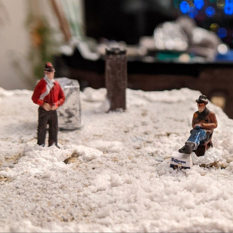 Two tiny guys sitting on a snowy flat rooftop. One is seated with his feet up on a case of Corona.